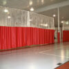 Indoor gym with red partition curtains and white safety netting, overhead lights, and a polished court floor with lines.