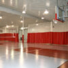 Indoor gym with a basketball hoop on the right and red curtain dividers separating courts under bright ceiling lights