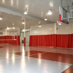 Indoor gym with a basketball hoop on the right and red curtain dividers separating courts under bright ceiling lights