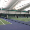 23955Z-1 | National Sports Products Indoor tennis center with players on a green court and a net across the foreground, blue surrounding floor visible for adjacent court.