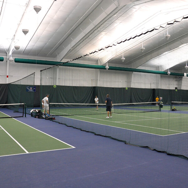 23955Z-1 | National Sports Products Indoor tennis center with players on a green court and a net across the foreground, blue surrounding floor visible for adjacent court.