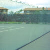 23955Z-2 | National Sports Products Tennis court enclosed by a green mesh fence, with a long net and white lines on the court; houses in the background under a cloudy sky.