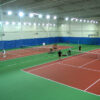 23955Z-4 | National Sports Products Indoor tennis facility with multiple courts, bright overhead lights, and players on red and green courts.