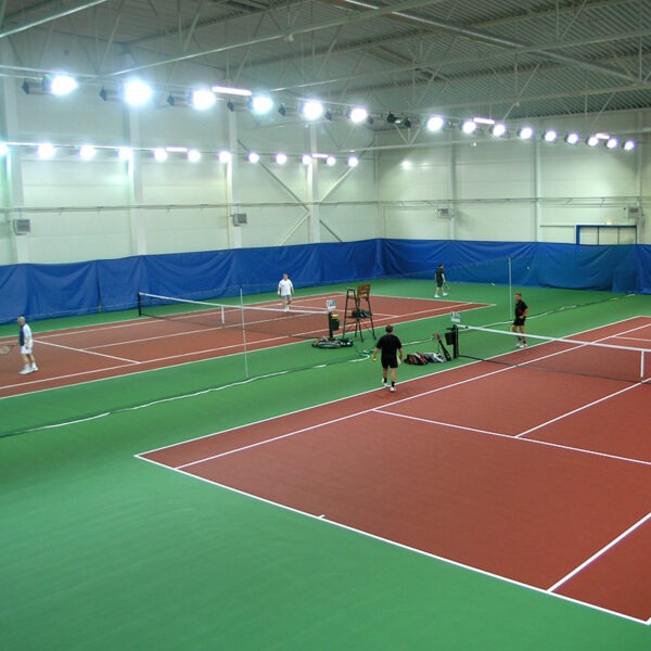 23955Z-4 | National Sports Products Indoor tennis facility with multiple courts, bright overhead lights, and players on red and green courts.