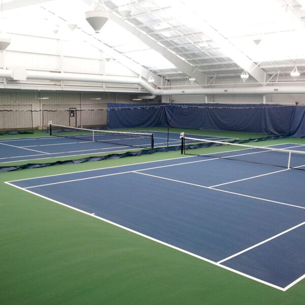 23955Z-5 | National Sports Products Interior of an indoor tennis facility with multiple blue courts, white boundary lines, and nets, surrounded by green flooring and bright overhead lights in a vaulted hall.