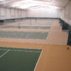 23955Z-7 | National Sports Products Indoor tennis facility with multiple green courts separated by nets and a brown floor under bright overhead lights.