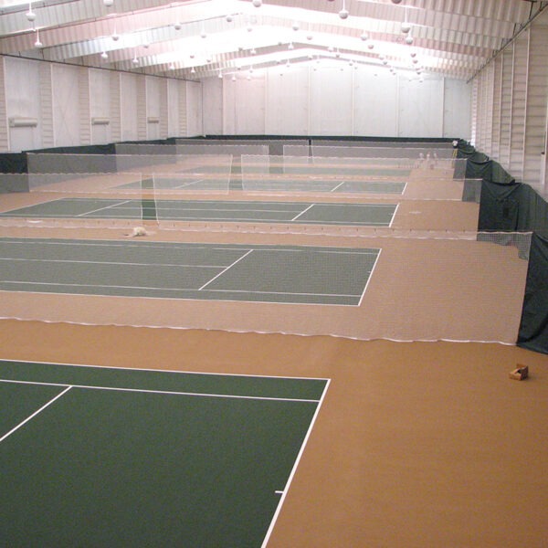 23955Z-7 | National Sports Products Indoor tennis facility with multiple green courts separated by nets and a brown floor under bright overhead lights.