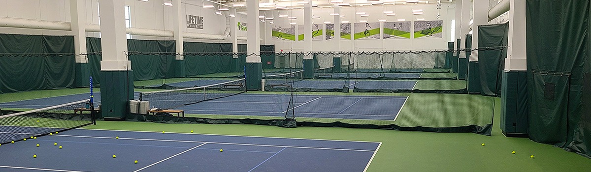 23955Z-a | National Sports Products Indoor sports facility with multiple blue courts, netted barriers, and scattered tennis balls on the green floor, banners on the walls.